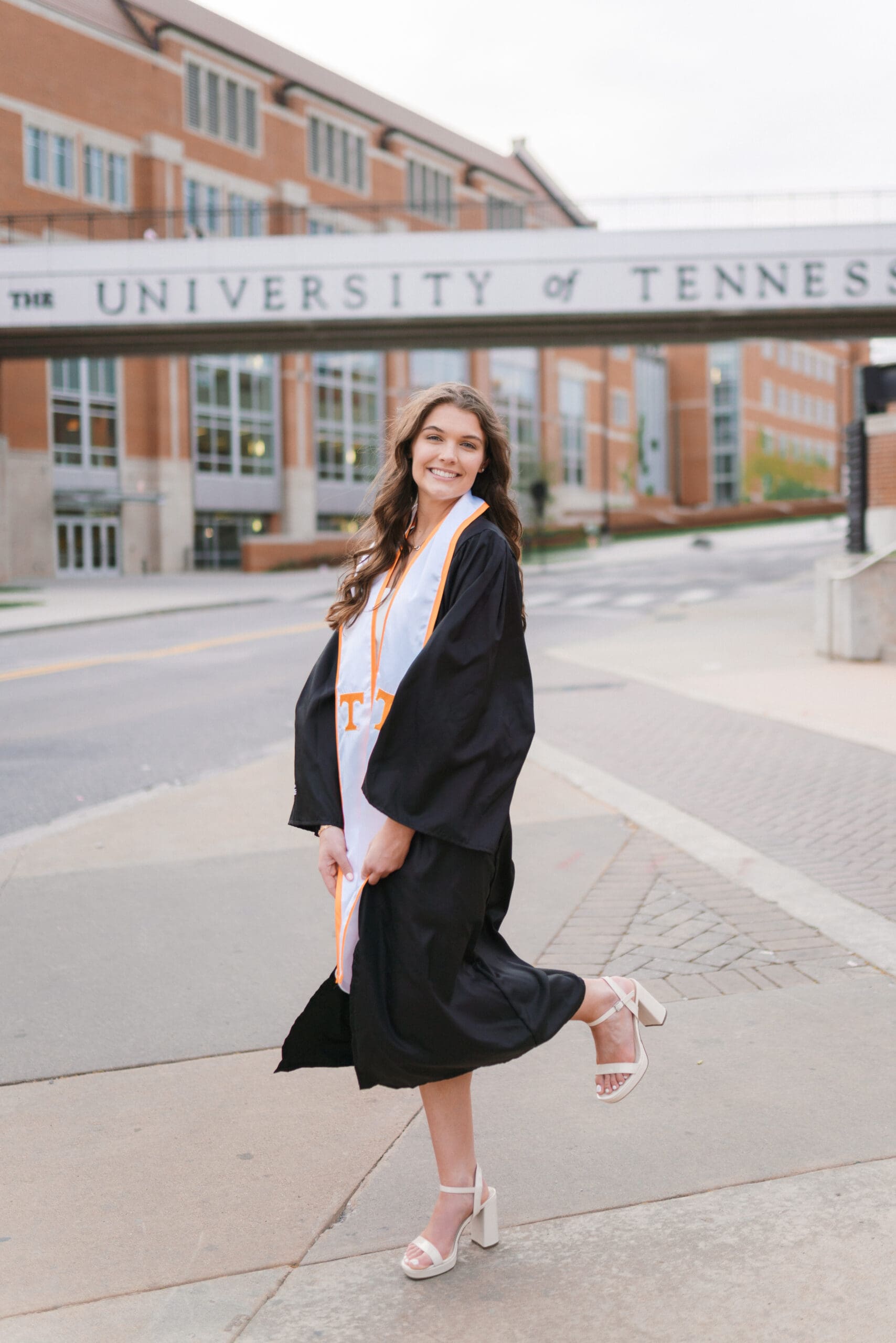 girl in graduation cap and gown stands in front of the university of tennessee knoxville bridge for senior photos