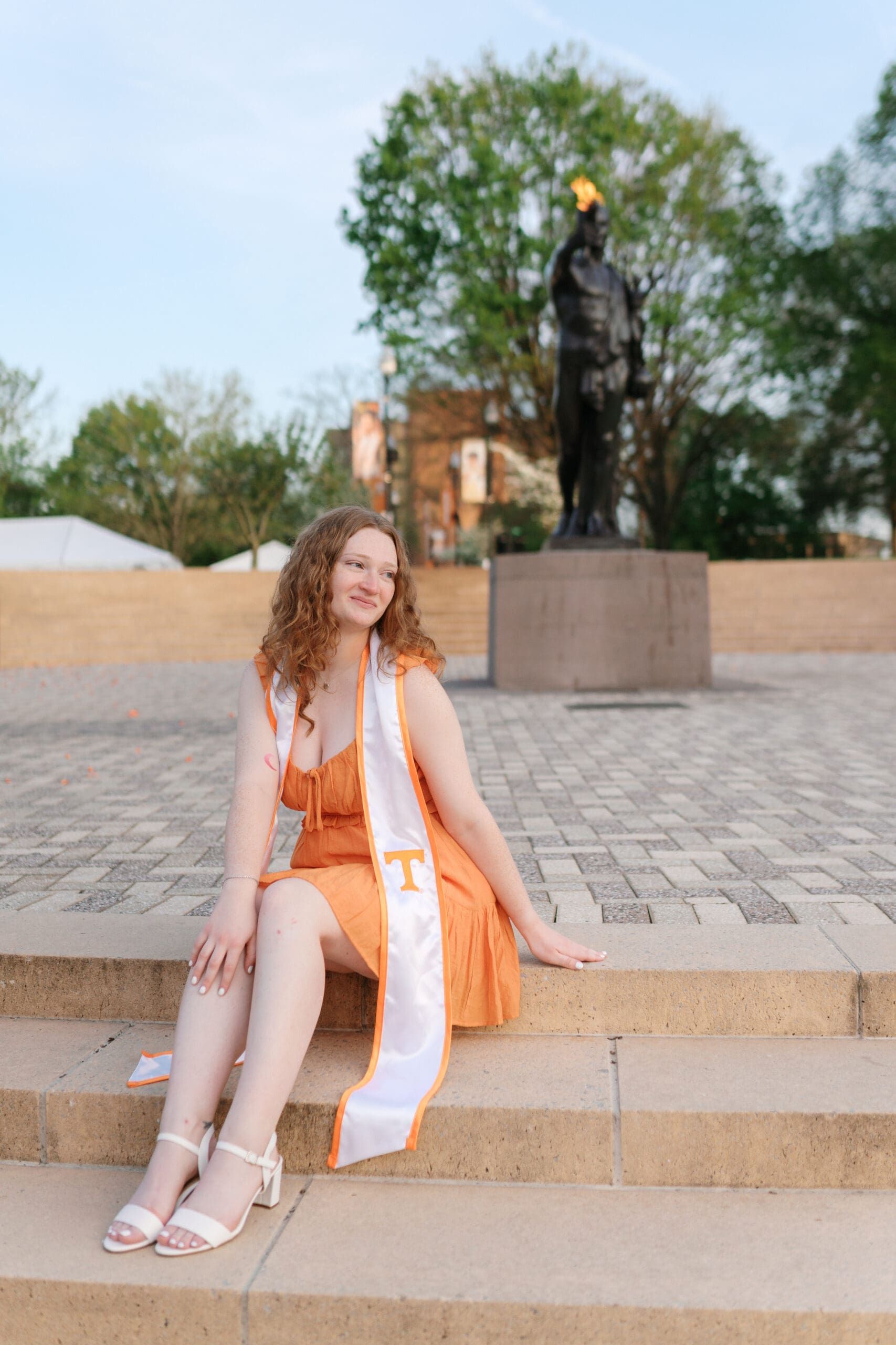senior in orange dress sits in front of the torchbearer statue during her graduation session at the university of tennessee knoxville